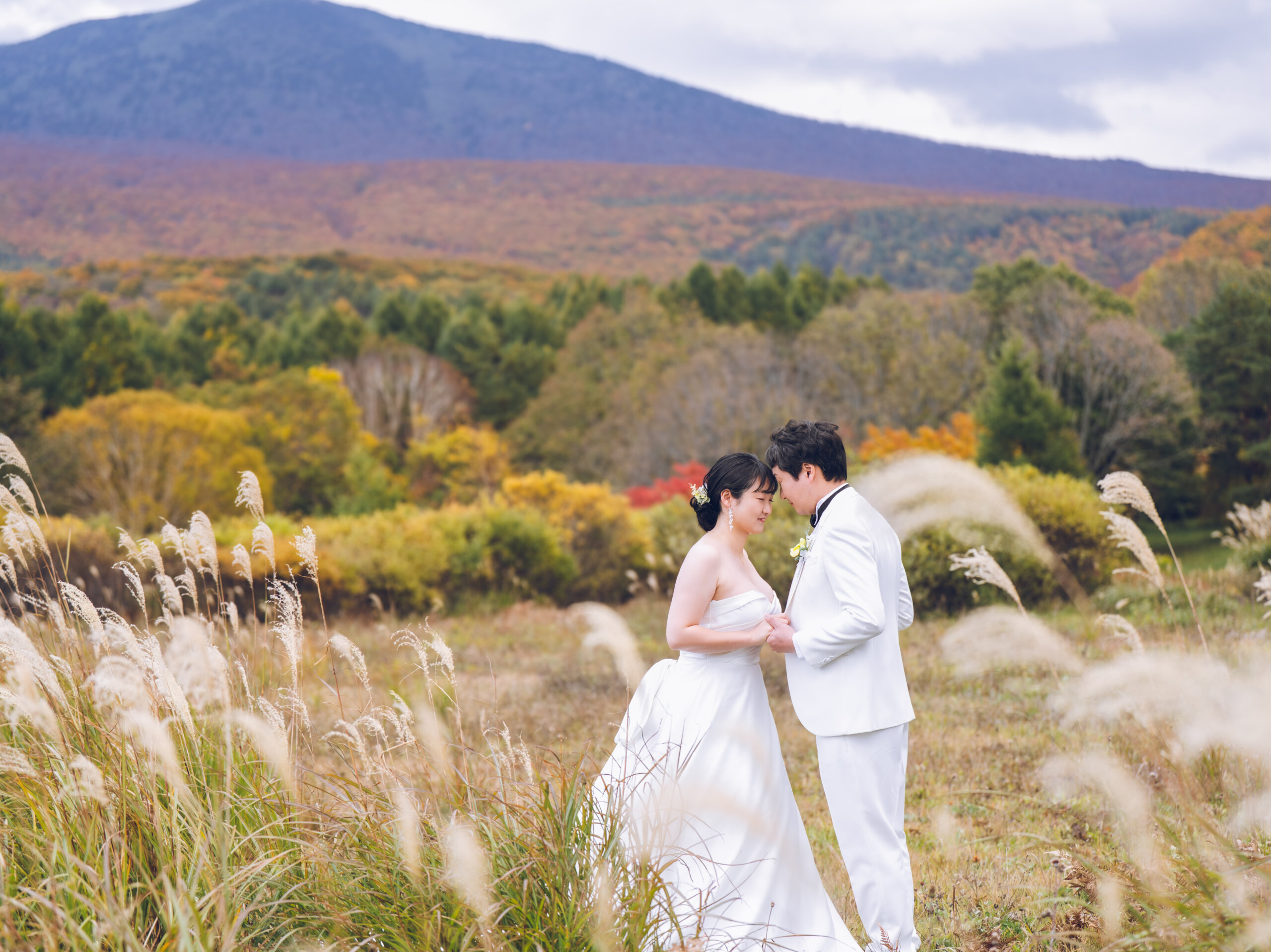 紅葉した山並みとススキを背景に寄り添う、白いドレスとタキシード姿の新郎新婦
