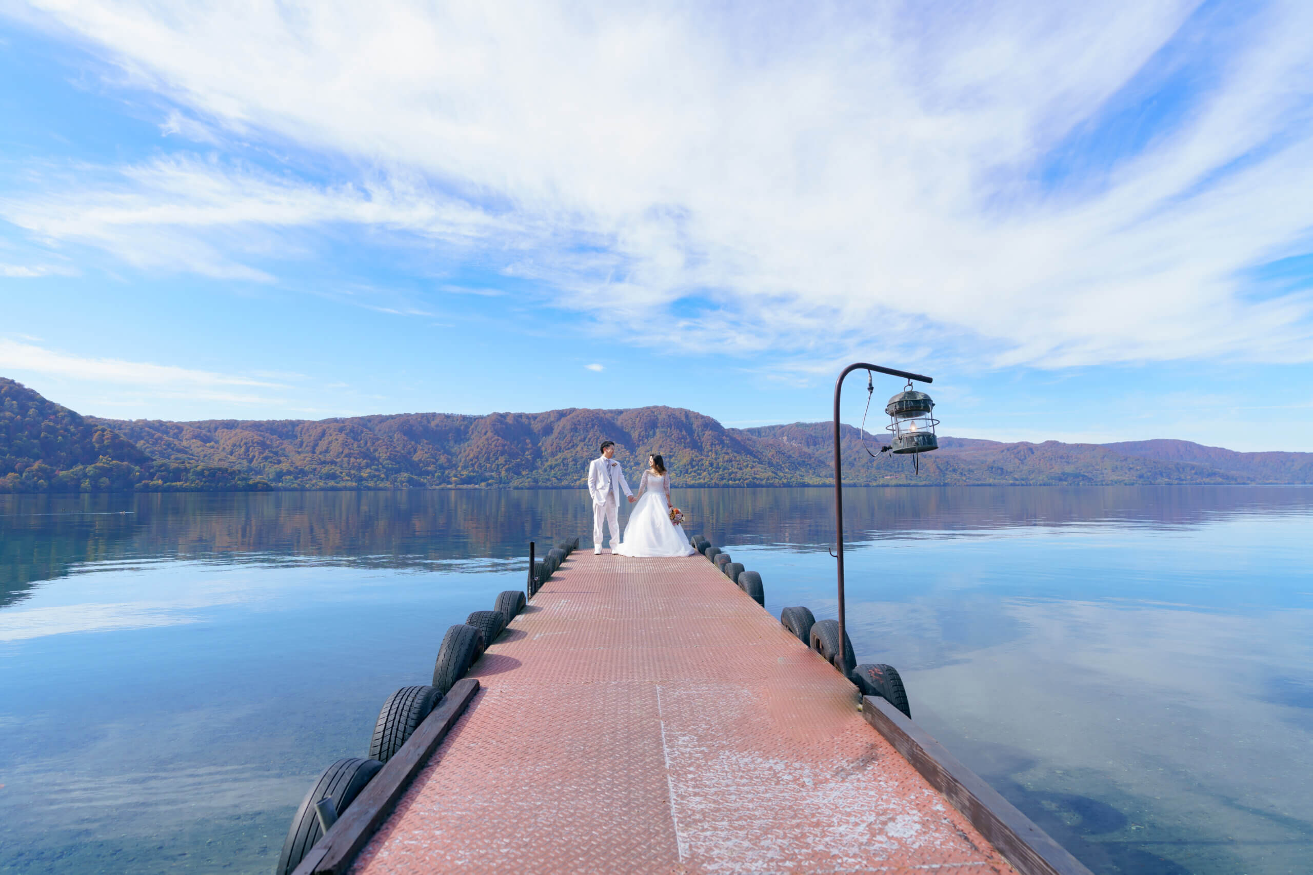 十和田湖の湖面と紅葉した山々を背景に、桟橋で手をつなぐ新郎新婦